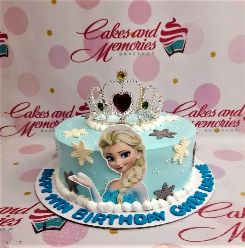 Light blue Frozen themed cake featuring Elsa printout, a silver rhinestone tiara, and white and gray fondant snowflakes. Light blue Frozen themed cake featuring Elsa printout, a silver rhinestone tiara, and white and gray fondant snowflakes.
