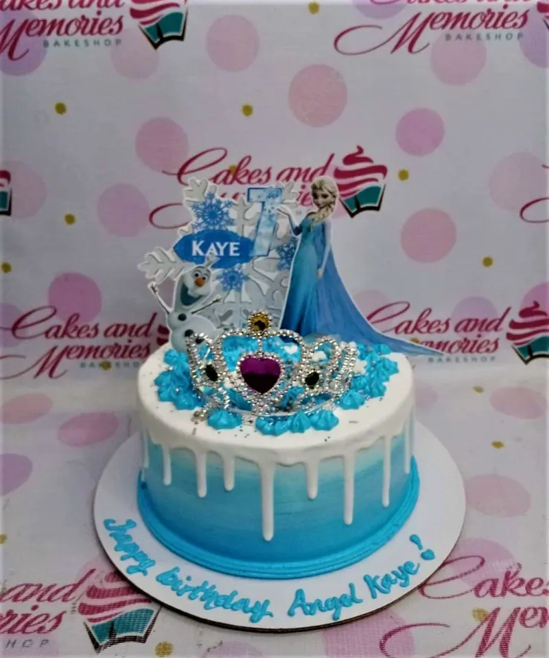 A blue, white, and silver 1 Tier cake themed around Frozen Elsa, featuring Elsa, Olaf, a silver tiara, and blue rosettes. A blue, white, and silver 1 Tier cake themed around Frozen Elsa, featuring Elsa, Olaf, a silver tiara, and blue rosettes.
