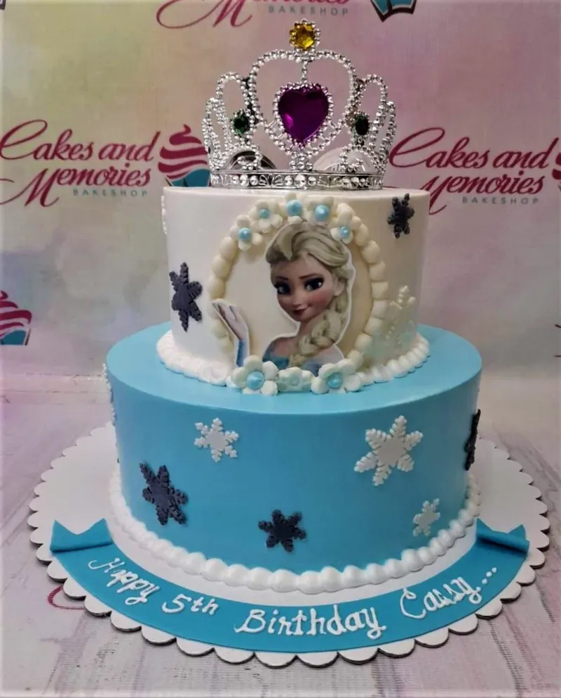 A blue and white two-tier cake themed around Frozen Elsa, featuring an Elsa printout, a silver tiara, and white and gray snowflakes. A blue and white two-tier cake themed around Frozen Elsa, featuring an Elsa printout, a silver tiara, and white and gray snowflakes.