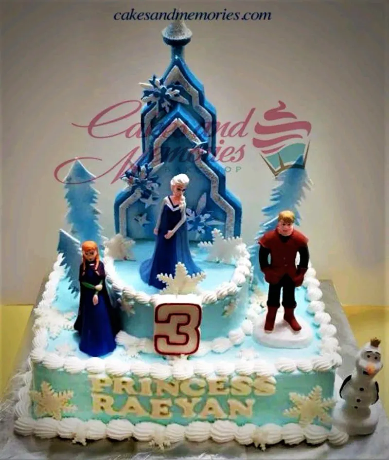 A blue and white 2-tier cake themed around Frozen, featuring Elsa, Anna, Kristoff, Olaf, a blue ice castle, and white rosettes. A blue and white 2-tier cake themed around Frozen, featuring Elsa, Anna, Kristoff, Olaf, a blue ice castle, and white rosettes.
