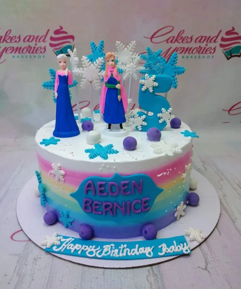 A blue, white, and rainbow gradient 1 Tier cake themed around Frozen, featuring Elsa and Anna figurines, a blue number five topper, white and blue snowflakes, and "AEDEN BERNICE" text. A blue, white, and rainbow gradient 1 Tier cake themed around Frozen, featuring Elsa and Anna figurines, a blue number five topper, white and blue snowflakes, and "AEDEN BERNICE" text.