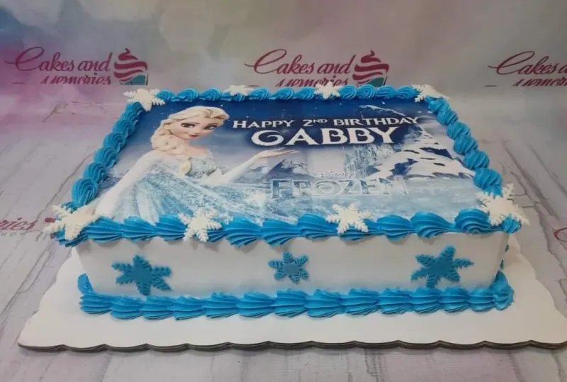 A white and blue Rectangle cake themed around Frozen, featuring an Elsa printout and white snowflakes, with blue icing borders. A white and blue Rectangle cake themed around Frozen, featuring an Elsa printout and white snowflakes, with blue icing borders.