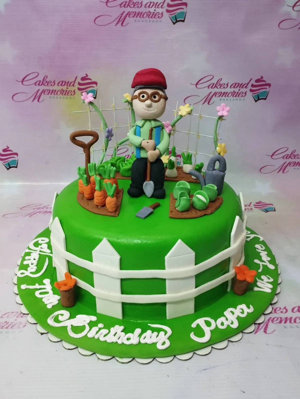A green 1 Tier Fondant garden cake, featuring a 3D gardener figure, fondant carrots, cabbages, and a white picket fence. A green 1 Tier Fondant garden cake, featuring a 3D gardener figure, fondant carrots, cabbages, and a white picket fence.