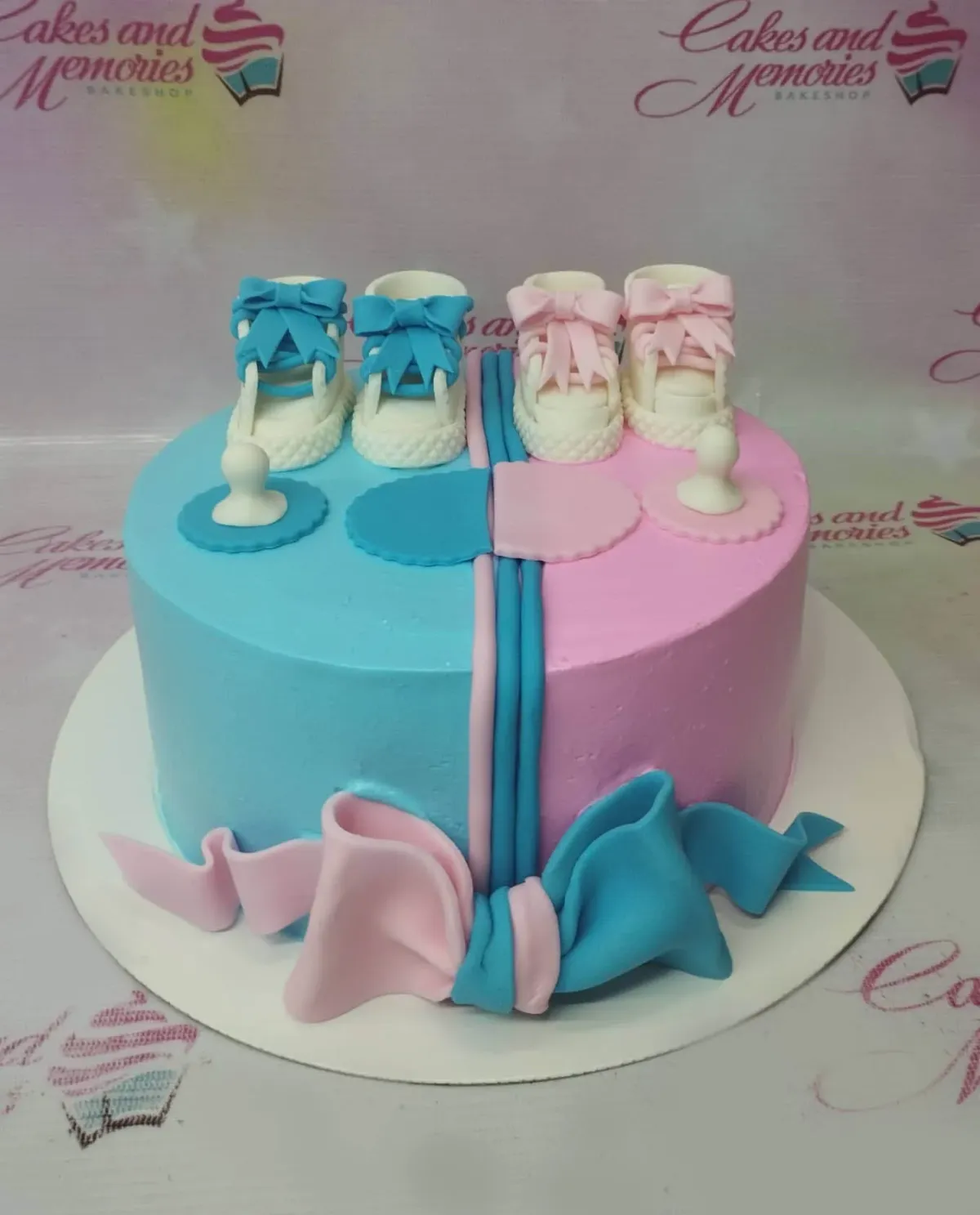 A blue and pink 1-tier cake themed around Gender Reveal, featuring fondant baby shoes, pacifiers, and a large bow at the base. A blue and pink 1-tier cake themed around Gender Reveal, featuring fondant baby shoes, pacifiers, and a large bow at the base.