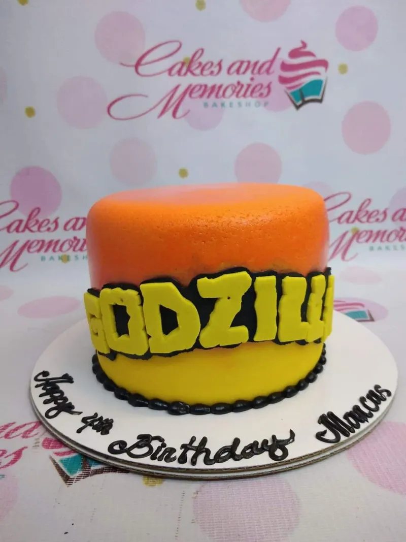 A orange and yellow single-tier Godzilla cake, featuring a yellow logo and 'Happy 4th Birthday Marcus' text. A orange and yellow single-tier Godzilla cake, featuring a yellow logo and 'Happy 4th Birthday Marcus' text.