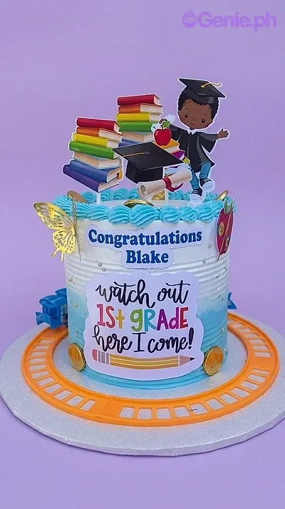 A blue and white 1 Tier cake themed around Graduation, featuring a student in a gown, school books, and a cap, with piped blue rosettes and a message panel. A blue and white 1 Tier cake themed around Graduation, featuring a student in a gown, school books, and a cap, with piped blue rosettes and a message panel.