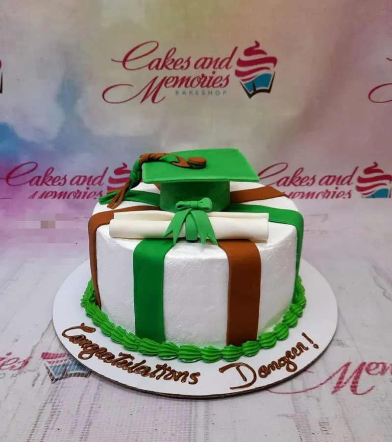 A white, green, and brown Graduation cake, featuring a green cap, white diploma, and ribbons. A white, green, and brown Graduation cake, featuring a green cap, white diploma, and ribbons.