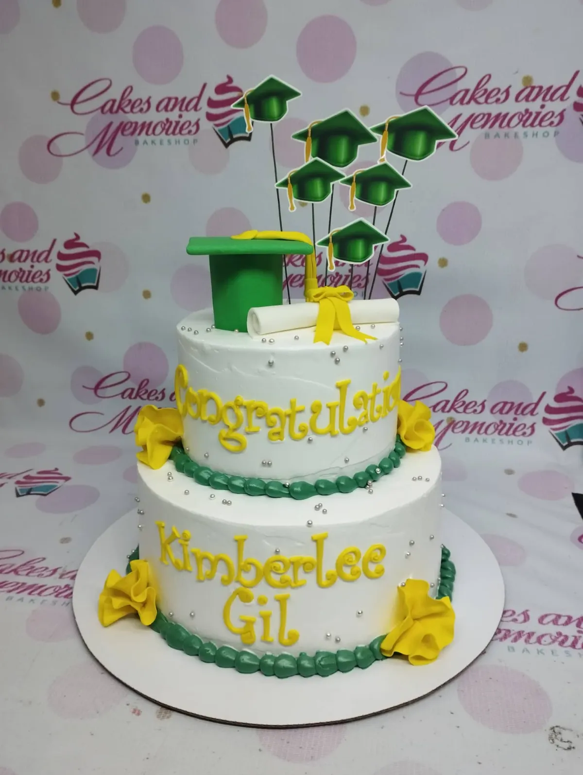 A white two-tier cake themed around Graduation, with a green mortarboard, white diploma, green cap printouts, yellow ruffles, and silver dragees. A white two-tier cake themed around Graduation, with a green mortarboard, white diploma, green cap printouts, yellow ruffles, and silver dragees.