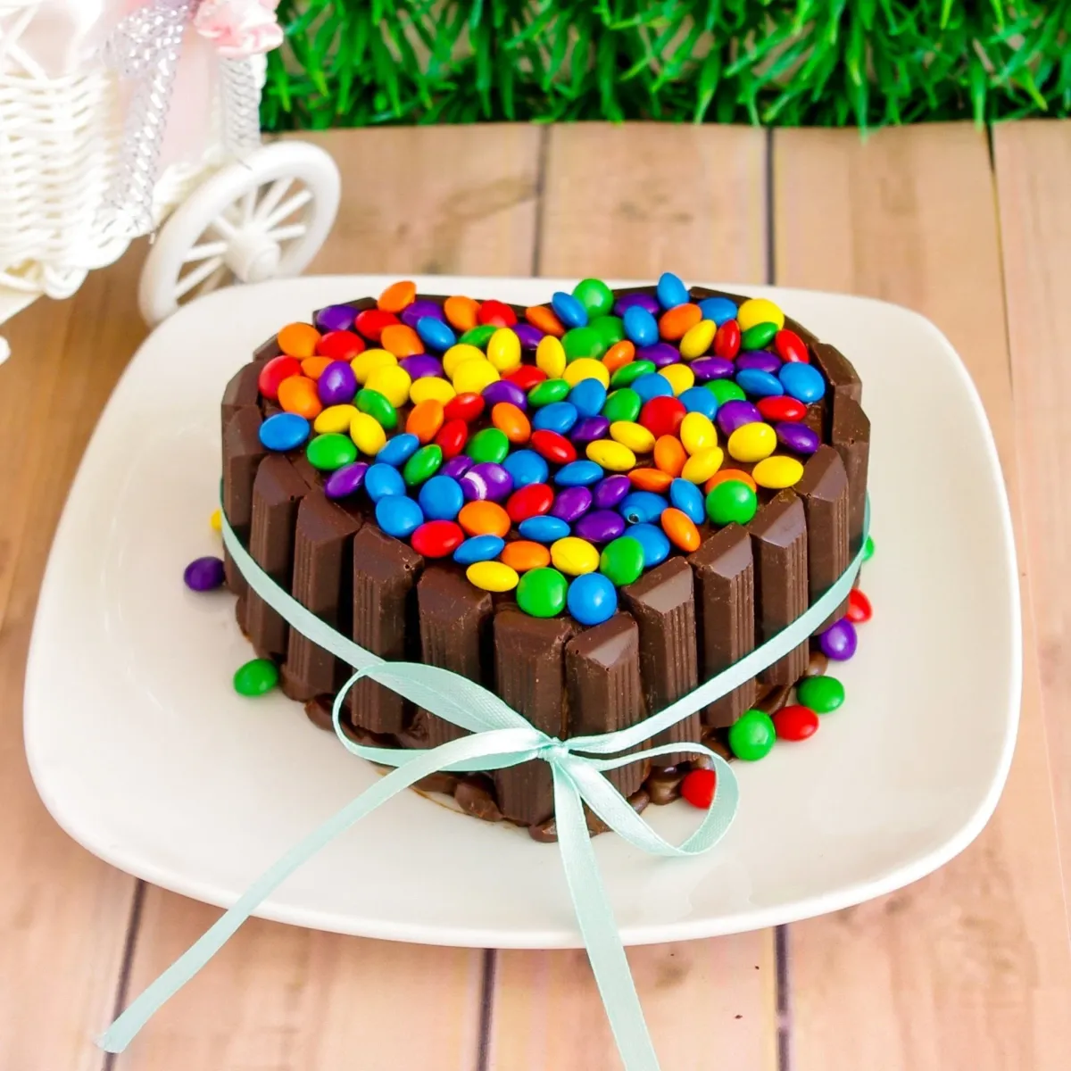 A brown heart-shaped 1-tier cake, topped with multi-colored chocolate candies, bordered by chocolate bars, and a blue ribbon. A brown heart-shaped 1-tier cake, topped with multi-colored chocolate candies, bordered by chocolate bars, and a blue ribbon.