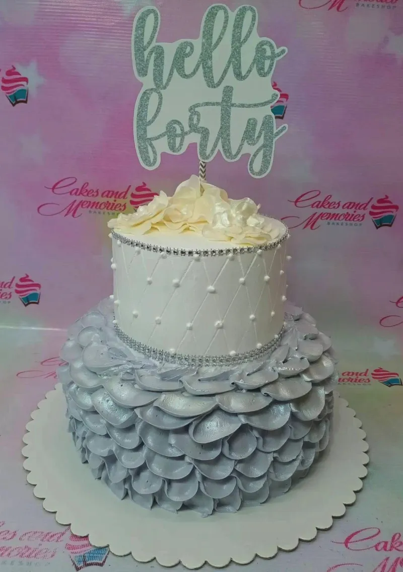 A white and silver two-tier cake themed around Hello Forty, featuring a silver 'Hello Forty' topper and silver ruffled icing. A white and silver two-tier cake themed around Hello Forty, featuring a silver 'Hello Forty' topper and silver ruffled icing.