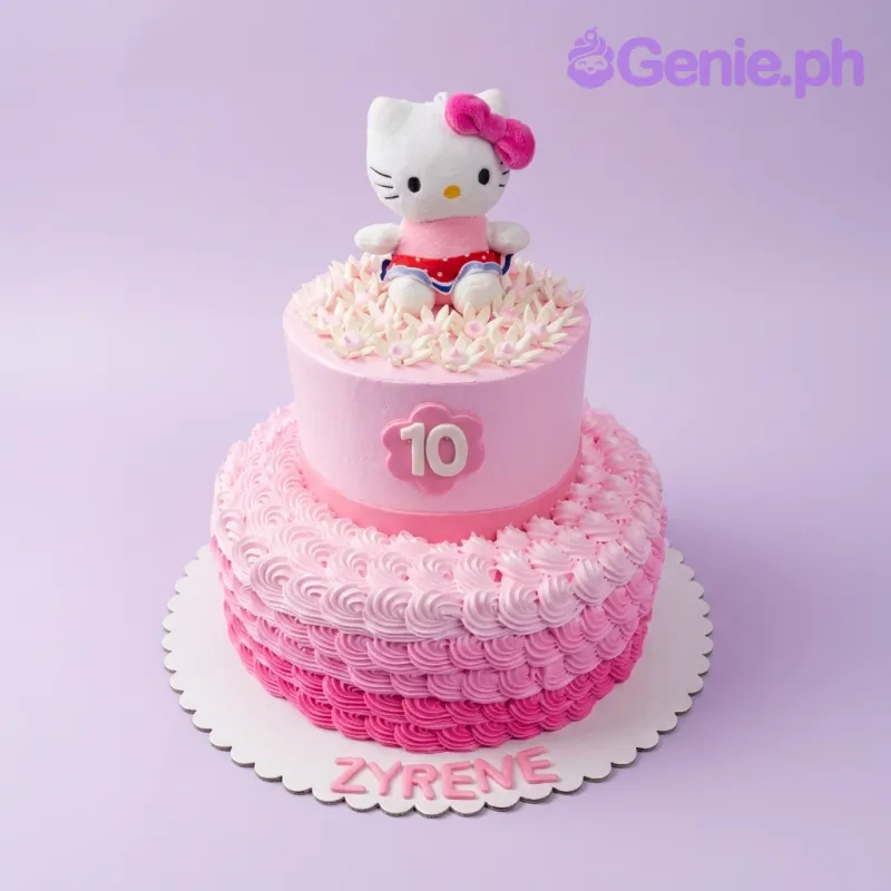 A pink and white 2-Tier cake themed around Hello Kitty, featuring a plush Hello Kitty topper and ombre icing rosettes with floral decorations. A pink and white 2-Tier cake themed around Hello Kitty, featuring a plush Hello Kitty topper and ombre icing rosettes with floral decorations.
