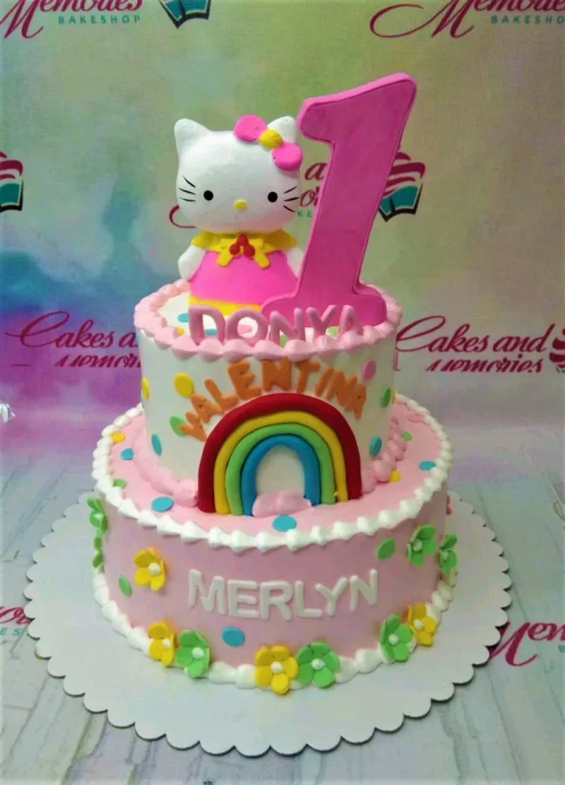 A pink and white two-tier cake themed Hello Kitty Rainbow, featuring a Hello Kitty figure, a pink number one, a rainbow, and edible flowers. A pink and white two-tier cake themed Hello Kitty Rainbow, featuring a Hello Kitty figure, a pink number one, a rainbow, and edible flowers.