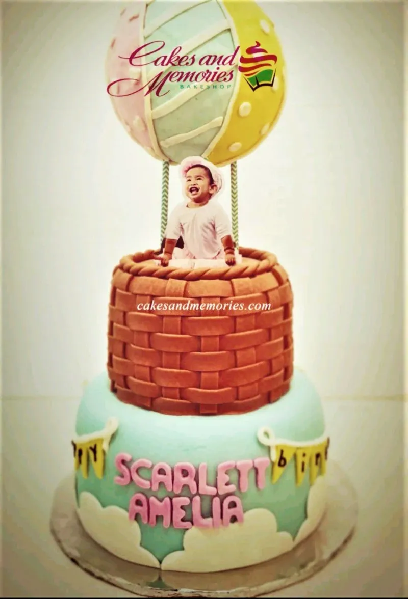 A multi-colored two-tier fondant Hot Air Balloon cake, featuring a hot air balloon, baby photo, basket-weave, and clouds. A multi-colored two-tier fondant Hot Air Balloon cake, featuring a hot air balloon, baby photo, basket-weave, and clouds.