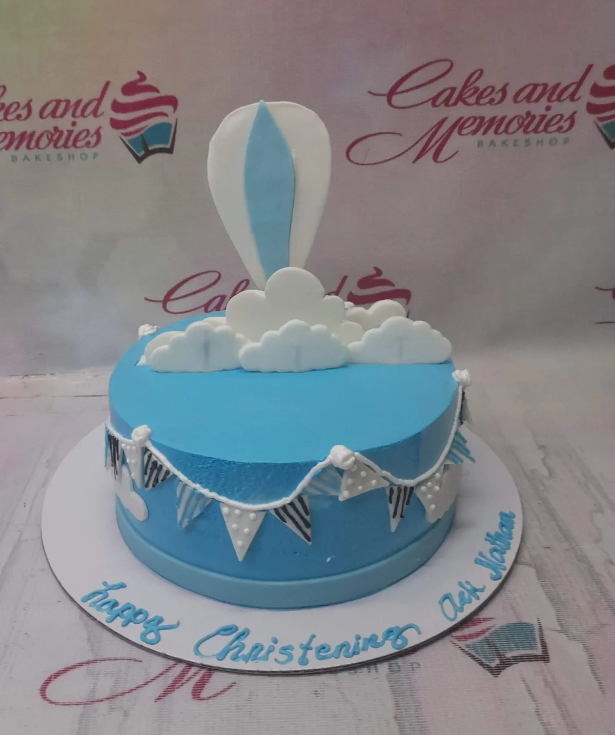 A light blue christening cake with a hot air balloon topper, white fondant clouds, and decorative bunting. A light blue christening cake with a hot air balloon topper, white fondant clouds, and decorative bunting.