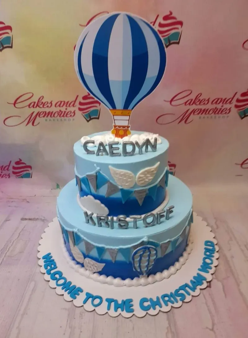 A two-tier light blue hot air balloon baptism cake with silver gumpaste letters and blue bunting flags. A two-tier light blue hot air balloon baptism cake with silver gumpaste letters and blue bunting flags.