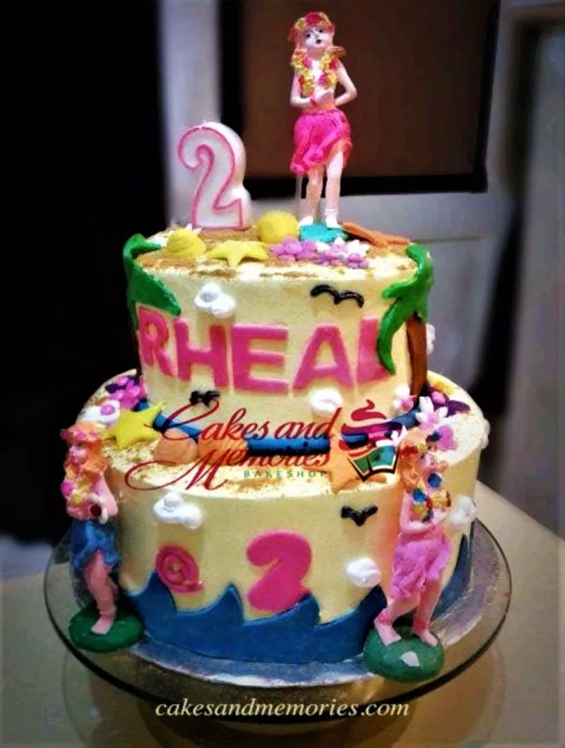 A yellow and pink two-tier cake themed Hula Girl Beach, with hula girl figurines, a number 2 candle, fondant letters, palm trees, and blue waves. A yellow and pink two-tier cake themed Hula Girl Beach, with hula girl figurines, a number 2 candle, fondant letters, palm trees, and blue waves.