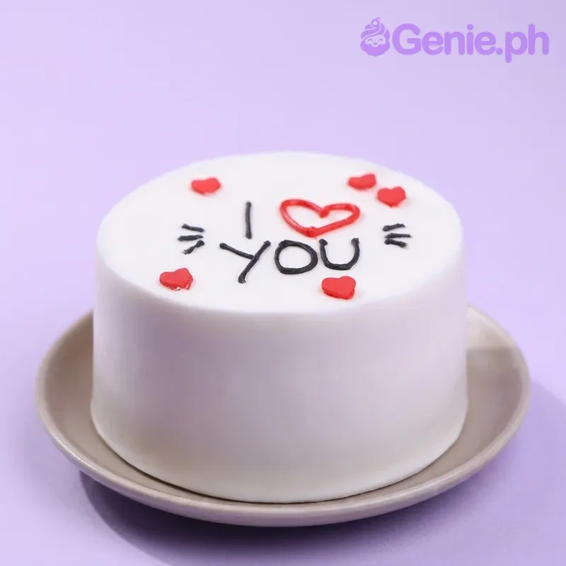 A white and red single tier cake with an 'I Heart You' theme, featuring red icing hearts and black piped text. A white and red single tier cake with an 'I Heart You' theme, featuring red icing hearts and black piped text.