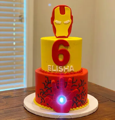 A yellow and red 2-tier cake themed around Iron Man, featuring an Iron Man mask, glowing arc reactor hand, and black icing doodles. A yellow and red 2-tier cake themed around Iron Man, featuring an Iron Man mask, glowing arc reactor hand, and black icing doodles.