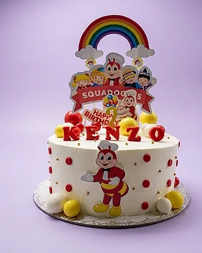 A white 1-tier Jollibee birthday cake with red polka dots, featuring Jollibee characters, a rainbow backdrop, a gold number 2 candle, and 'KENZO' lettering. A white 1-tier Jollibee birthday cake with red polka dots, featuring Jollibee characters, a rainbow backdrop, a gold number 2 candle, and 'KENZO' lettering.