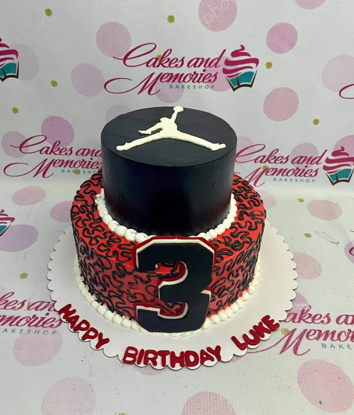 Two-tier Jordan themed birthday cake in black and red with a white Jumpman logo and a large number 3 on the side. Two-tier Jordan themed birthday cake in black and red with a white Jumpman logo and a large number 3 on the side.