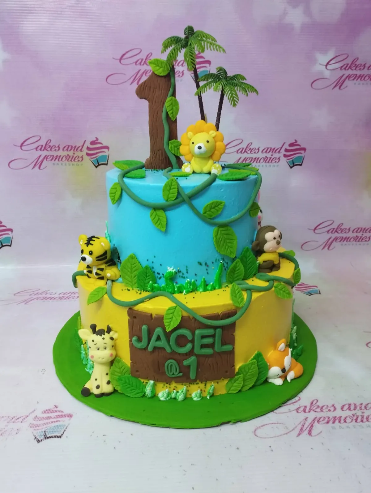 A blue and yellow two-tier Jungle Safari cake, with fondant lion, tiger, giraffe, monkey, fox, number one, green vines, and 'JACEL @ 1'. A blue and yellow two-tier Jungle Safari cake, with fondant lion, tiger, giraffe, monkey, fox, number one, green vines, and 'JACEL @ 1'.