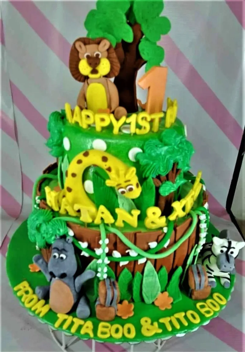 A green 3 Tier Fondant cake themed around Jungle Safari, featuring lion, giraffe, hippo, and zebra figures, with foliage and yellow lettering. A green 3 Tier Fondant cake themed around Jungle Safari, featuring lion, giraffe, hippo, and zebra figures, with foliage and yellow lettering.