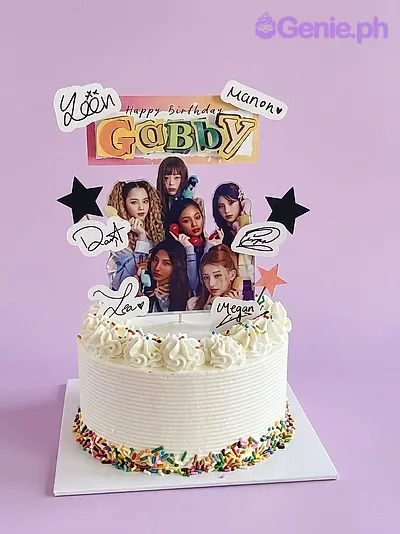 White soft-icing cake with colorful sprinkles at the base and multiple K-pop group printout toppers including a large Gabby name banner. White soft-icing cake with colorful sprinkles at the base and multiple K-pop group printout toppers including a large Gabby name banner.