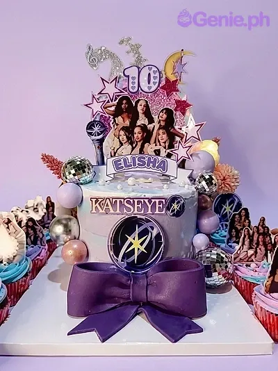A white and light blue 1-tier Katseye Kpop cake with group printouts, a circular logo, disco balls, and a large purple bow. A white and light blue 1-tier Katseye Kpop cake with group printouts, a circular logo, disco balls, and a large purple bow.