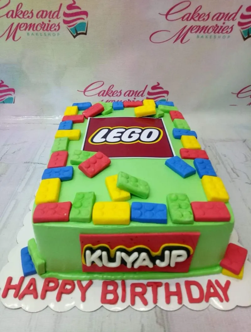 A green Lego-themed rectangle cake with a Lego logo printout, colorful fondant bricks, and personalized text. A green Lego-themed rectangle cake with a Lego logo printout, colorful fondant bricks, and personalized text.
