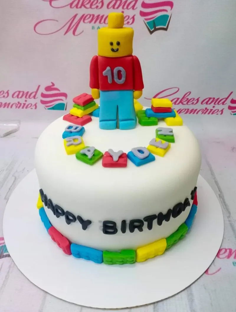 A white 1-tier fondant cake themed around Lego, featuring a large Lego man topper and colorful Lego brick decorations. A white 1-tier fondant cake themed around Lego, featuring a large Lego man topper and colorful Lego brick decorations.
