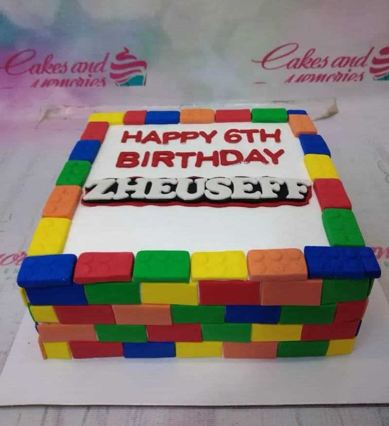A white square Lego-themed cake, featuring a personalized name topper and colorful fondant brick borders. A white square Lego-themed cake, featuring a personalized name topper and colorful fondant brick borders.