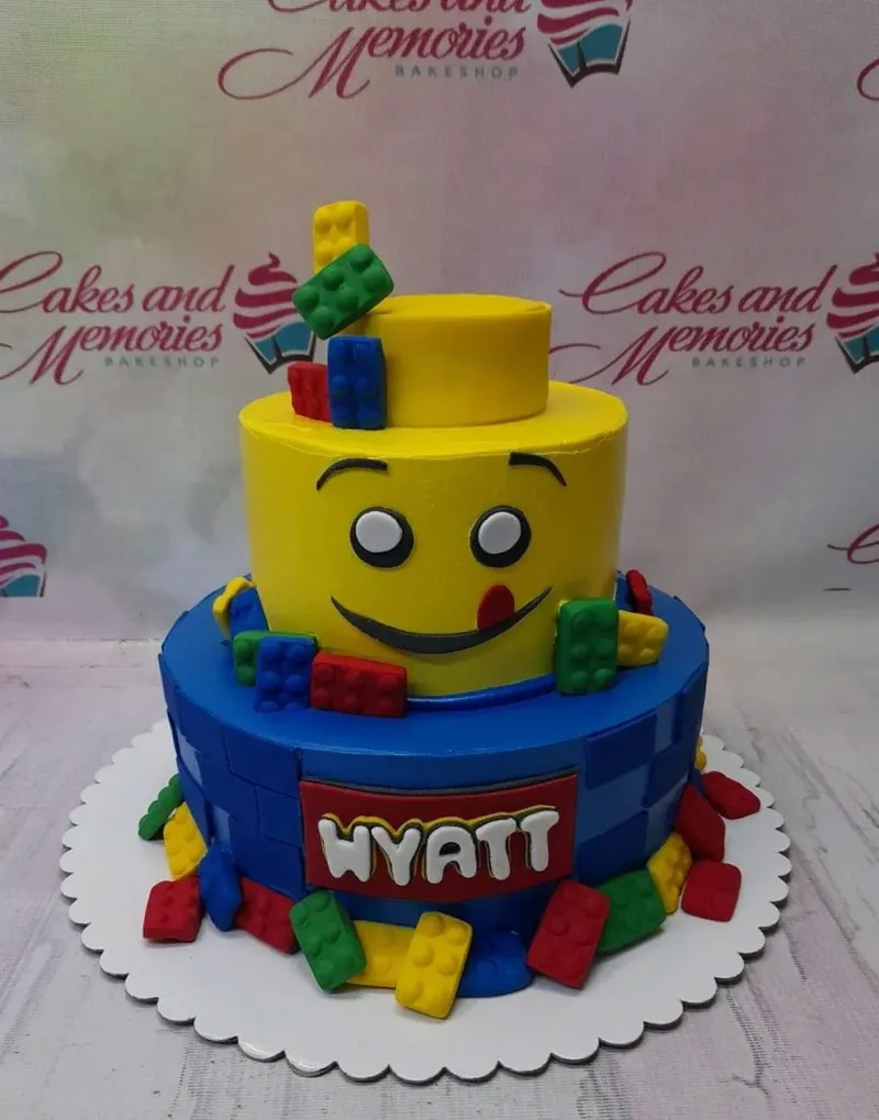 A yellow and blue two-tier fondant cake with a Lego theme, featuring a yellow Lego head and colorful brick accents. A yellow and blue two-tier fondant cake with a Lego theme, featuring a yellow Lego head and colorful brick accents.