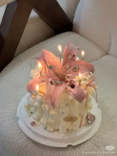 White 1-tier cake with soft icing, a large pink lily flower topper, and tall lit yellow birthday candles White 1-tier cake with soft icing, a large pink lily flower topper, and tall lit yellow birthday candles