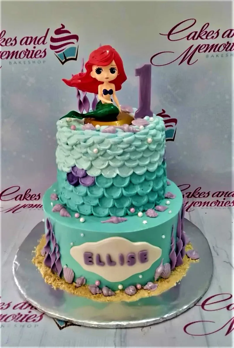 A vibrant two-tier Little Mermaid cake, featuring an Ariel figure, number 1 candle, scales, and shells. A vibrant two-tier Little Mermaid cake, featuring an Ariel figure, number 1 candle, scales, and shells.