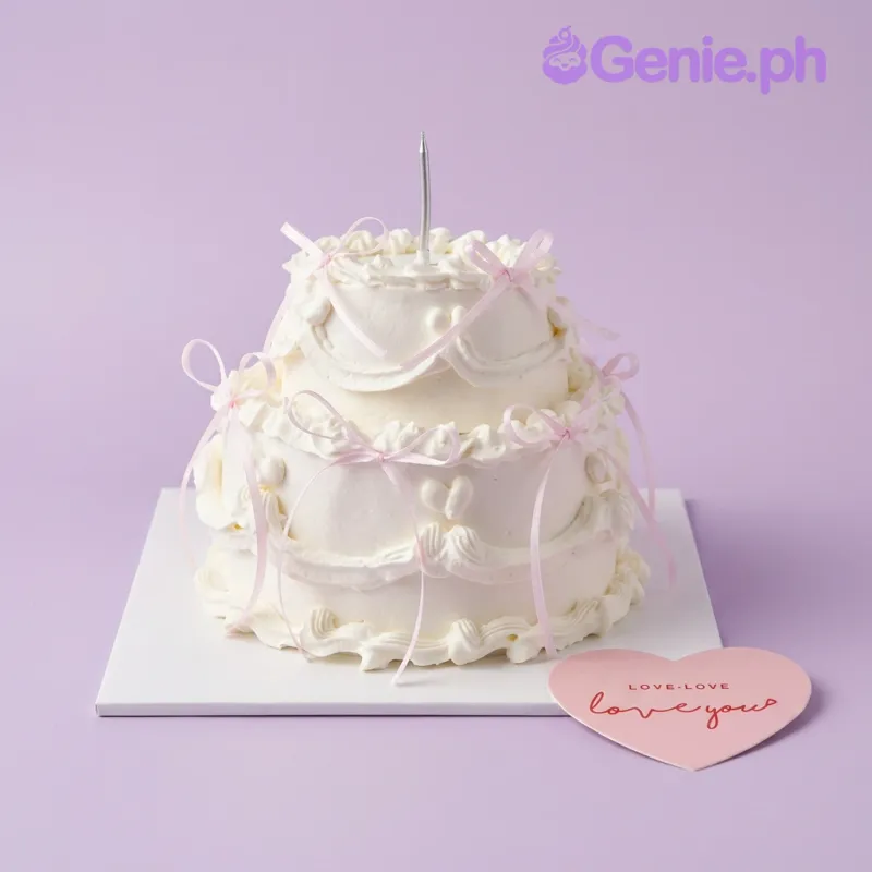 Two tier white soft iced cake with a medium pink heart printout topper and small pink satin ribbon bows Two tier white soft iced cake with a medium pink heart printout topper and small pink satin ribbon bows