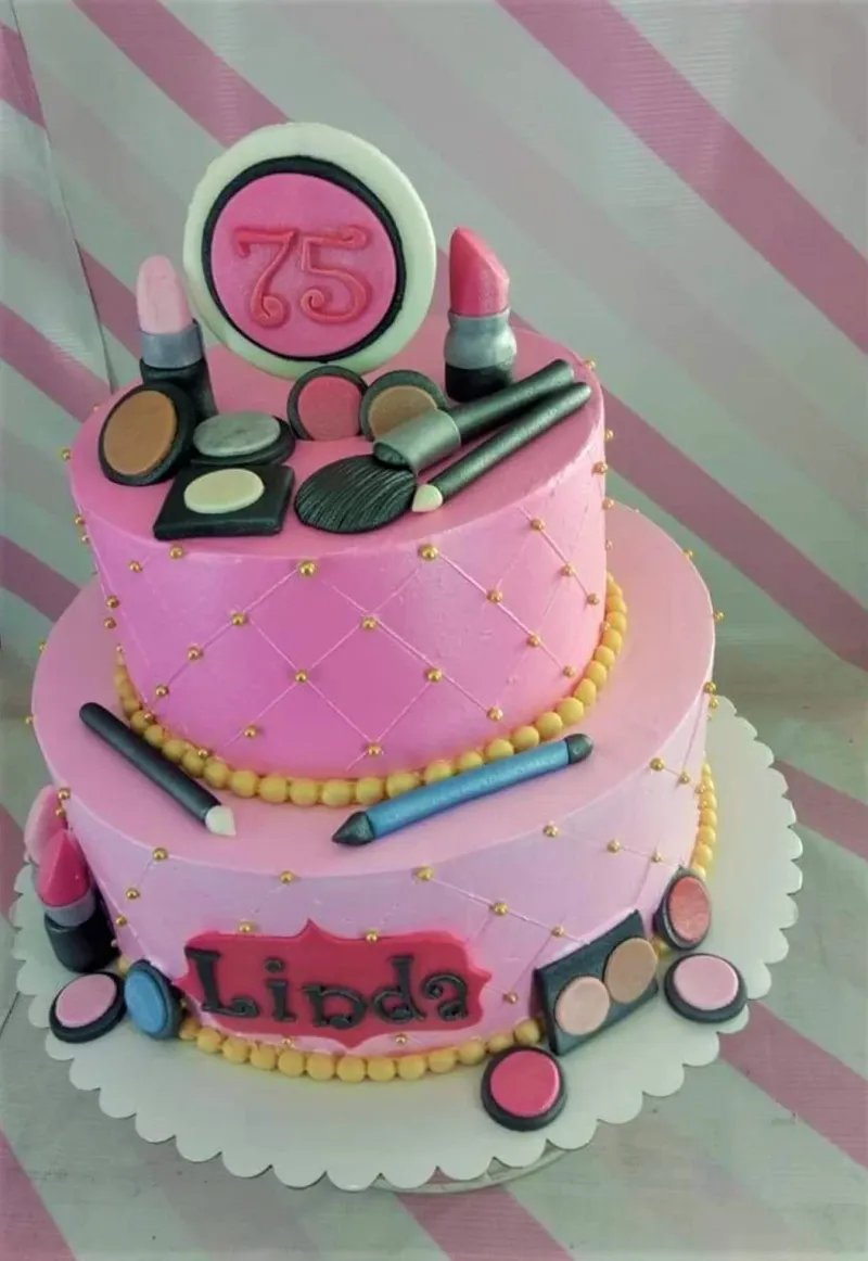 A pink 2-tier cake themed around makeup, featuring fondant lipsticks, brushes, a '75' plaque, and makeup compacts. A pink 2-tier cake themed around makeup, featuring fondant lipsticks, brushes, a '75' plaque, and makeup compacts.