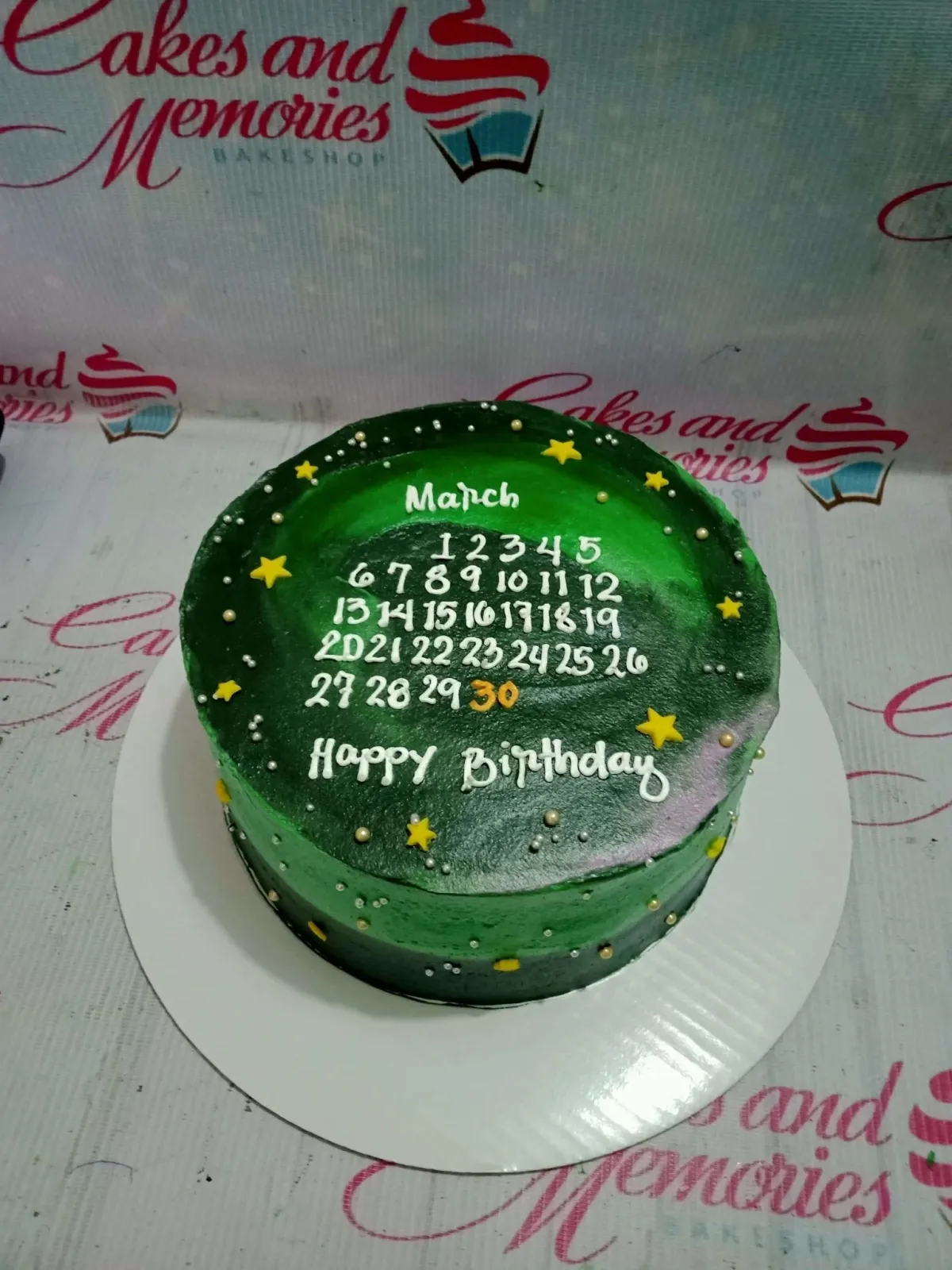A green 1-Tier cake themed as a March Calendar, featuring white icing text, yellow stars, silver dragees, and an orange highlighted date. A green 1-Tier cake themed as a March Calendar, featuring white icing text, yellow stars, silver dragees, and an orange highlighted date.