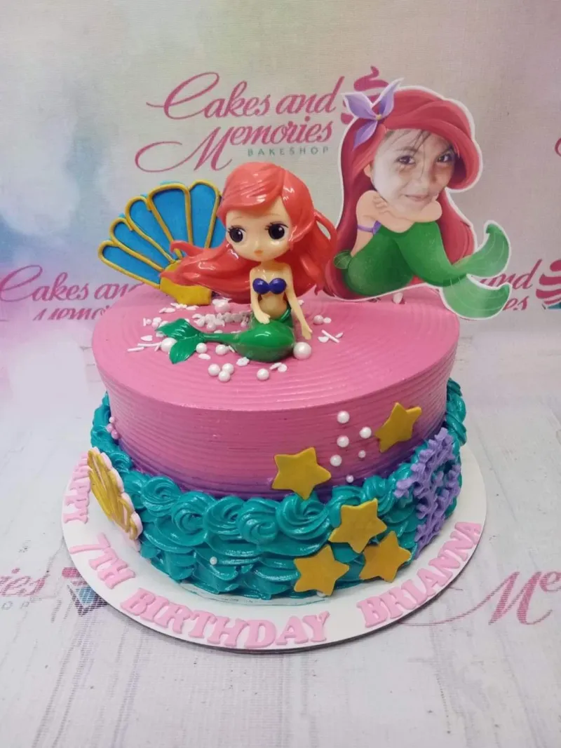 A pink and teal 1-tier mermaid-themed cake, featuring a 3D mermaid figure, a personalized photo topper, and teal icing ruffles with yellow stars. A pink and teal 1-tier mermaid-themed cake, featuring a 3D mermaid figure, a personalized photo topper, and teal icing ruffles with yellow stars.