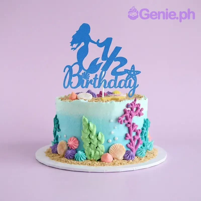 A blue 1-tier cake themed around Mermaid, featuring a blue glitter mermaid 1/2 birthday topper, edible sand, and gumpaste seashells. A blue 1-tier cake themed around Mermaid, featuring a blue glitter mermaid 1/2 birthday topper, edible sand, and gumpaste seashells.