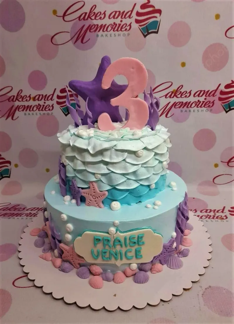 Two-tier mermaid themed cake with light blue and teal icing scales, purple starfish, and a large pink number three topper. Two-tier mermaid themed cake with light blue and teal icing scales, purple starfish, and a large pink number three topper.