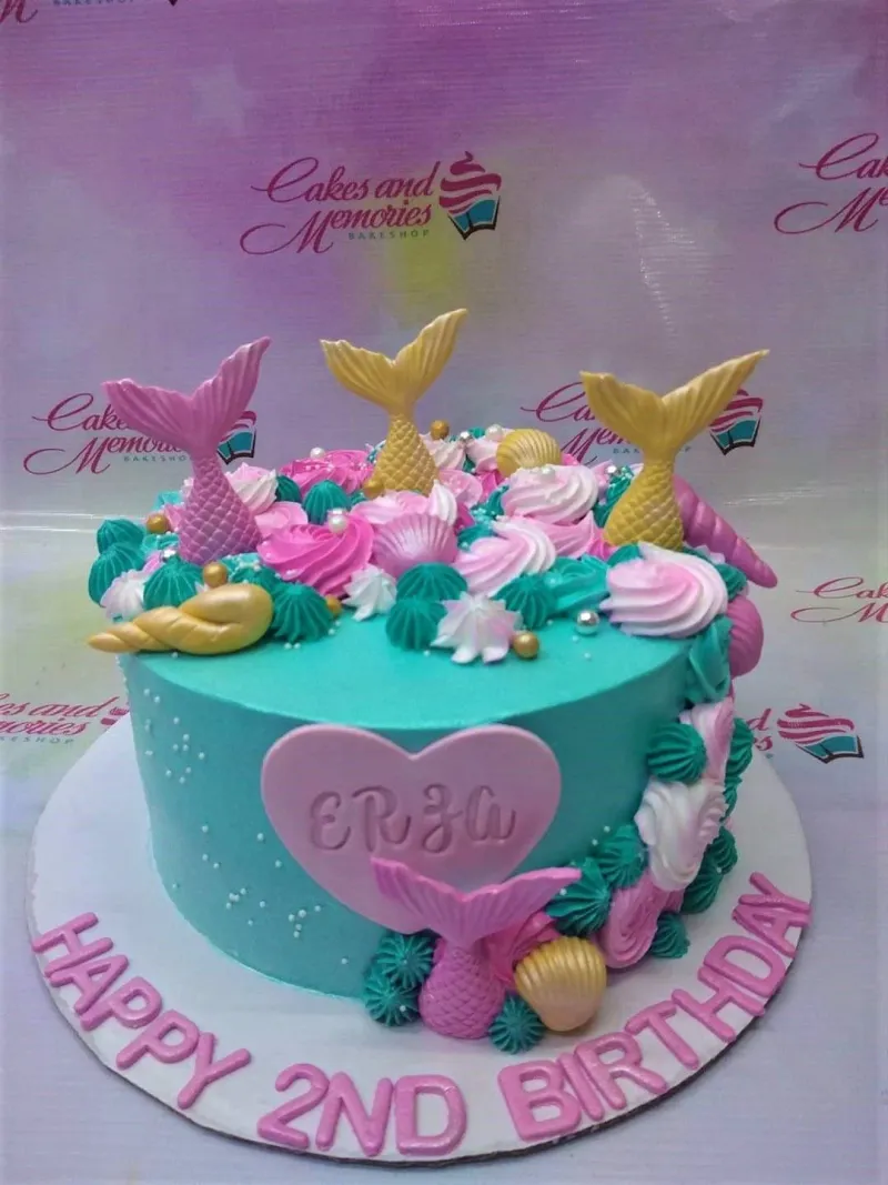 A teal single-tier cake themed around mermaids, featuring pink and gold mermaid tails, a pink heart name plaque, and various icing rosettes. A teal single-tier cake themed around mermaids, featuring pink and gold mermaid tails, a pink heart name plaque, and various icing rosettes.