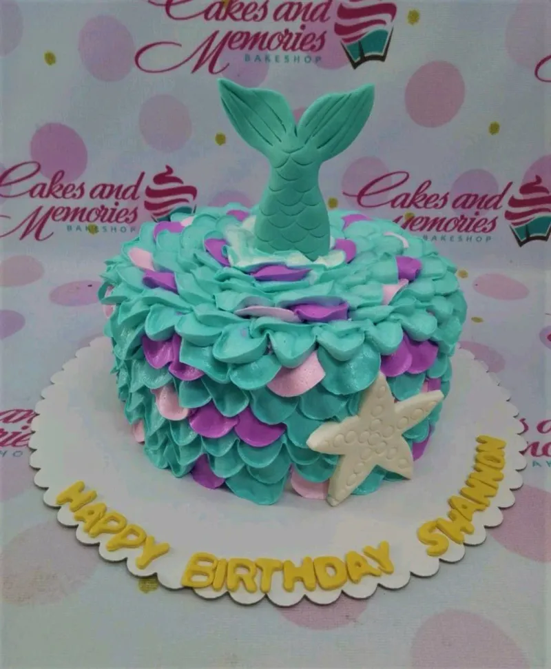 A multicolored 1-tier cake themed Mermaid, featuring a teal mermaid tail, white starfish, and scale-like icing. A multicolored 1-tier cake themed Mermaid, featuring a teal mermaid tail, white starfish, and scale-like icing.