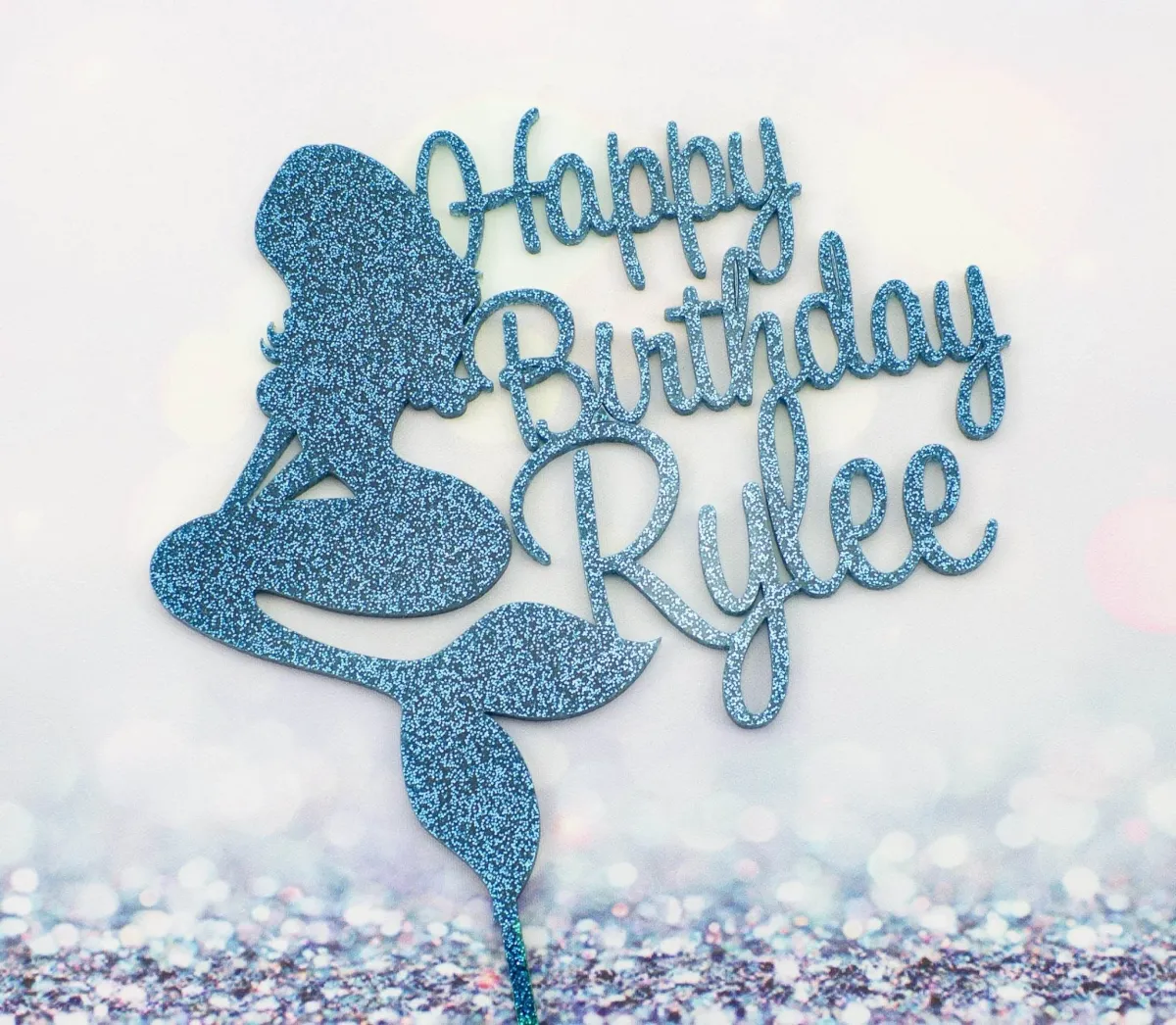 A white and blue 1-tier cake themed around Mermaid, featuring a blue glitter mermaid silhouette and 'Happy Birthday Rylee' text. A white and blue 1-tier cake themed around Mermaid, featuring a blue glitter mermaid silhouette and 'Happy Birthday Rylee' text.