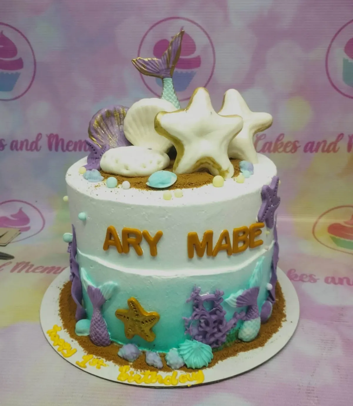 A white, teal, purple, and gold 1-Tier cake themed around Mermaid, featuring a mermaid tail, starfish, seashells, and personalized messages. A white, teal, purple, and gold 1-Tier cake themed around Mermaid, featuring a mermaid tail, starfish, seashells, and personalized messages.