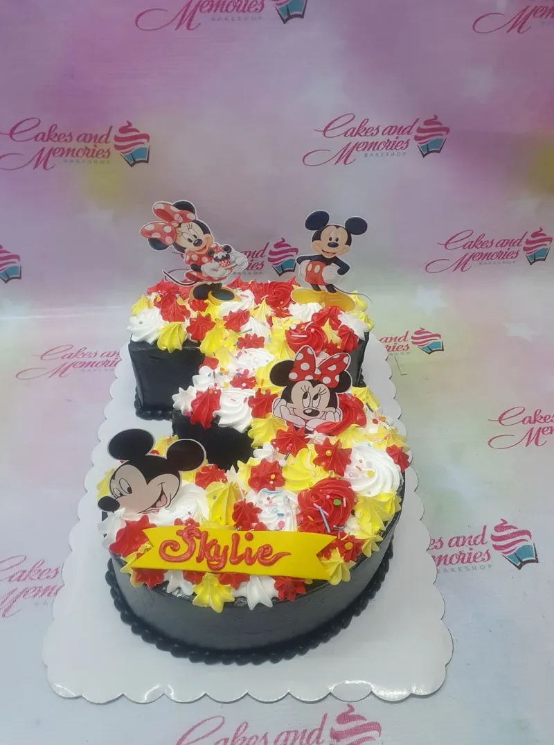 A black rectangle Mickey Minnie Mouse cake, featuring character printouts, a yellow name banner, icing rosettes, and sprinkles. A black rectangle Mickey Minnie Mouse cake, featuring character printouts, a yellow name banner, icing rosettes, and sprinkles.