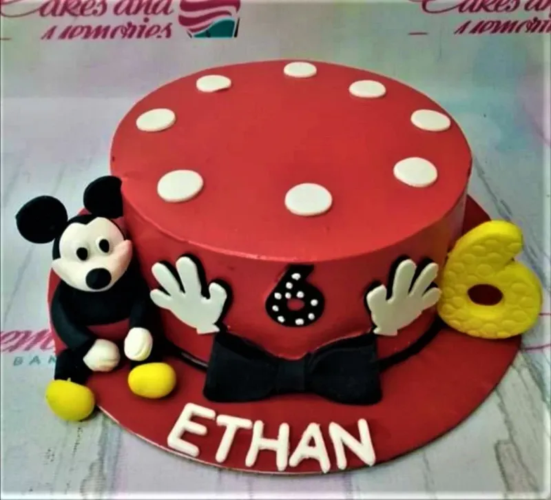 A red, white, and black 1-tier Mickey Mouse themed cake, featuring a 3D Mickey Mouse figure, yellow number six, white polka dots, and a black bow tie. A red, white, and black 1-tier Mickey Mouse themed cake, featuring a 3D Mickey Mouse figure, yellow number six, white polka dots, and a black bow tie.