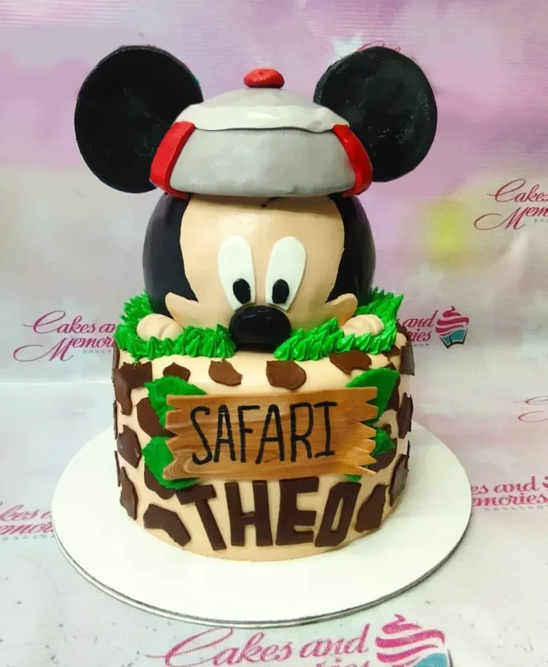 A beige and brown 1 Tier Fondant cake, Mickey Mouse Safari themed, with a 3D Mickey head in a safari hat, giraffe print, and 'SAFARI THEO' text. A beige and brown 1 Tier Fondant cake, Mickey Mouse Safari themed, with a 3D Mickey head in a safari hat, giraffe print, and 'SAFARI THEO' text.