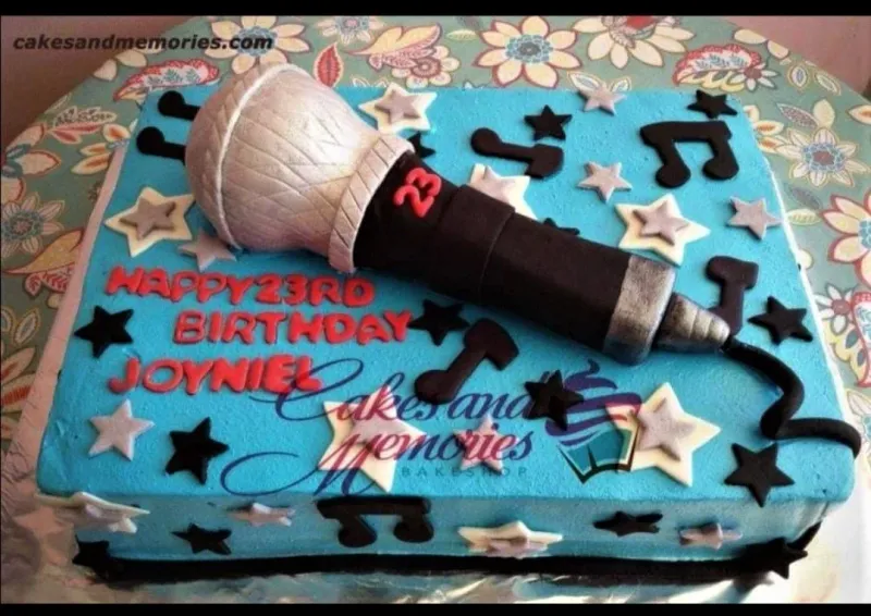A blue rectangular music-themed cake, featuring a 3D microphone, black music notes, silver stars, and red birthday text. A blue rectangular music-themed cake, featuring a 3D microphone, black music notes, silver stars, and red birthday text.