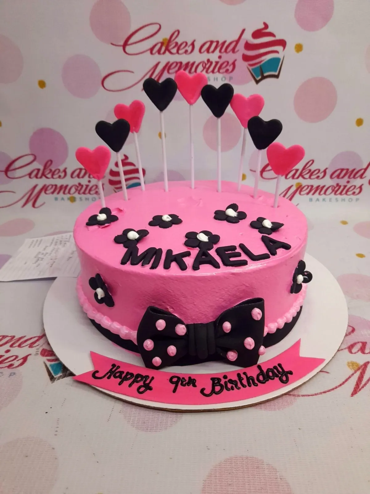 A pink 1-tier birthday cake for Mikaela, with black and pink heart toppers, black fondant flowers, and a black polka dot bow. A pink 1-tier birthday cake for Mikaela, with black and pink heart toppers, black fondant flowers, and a black polka dot bow.