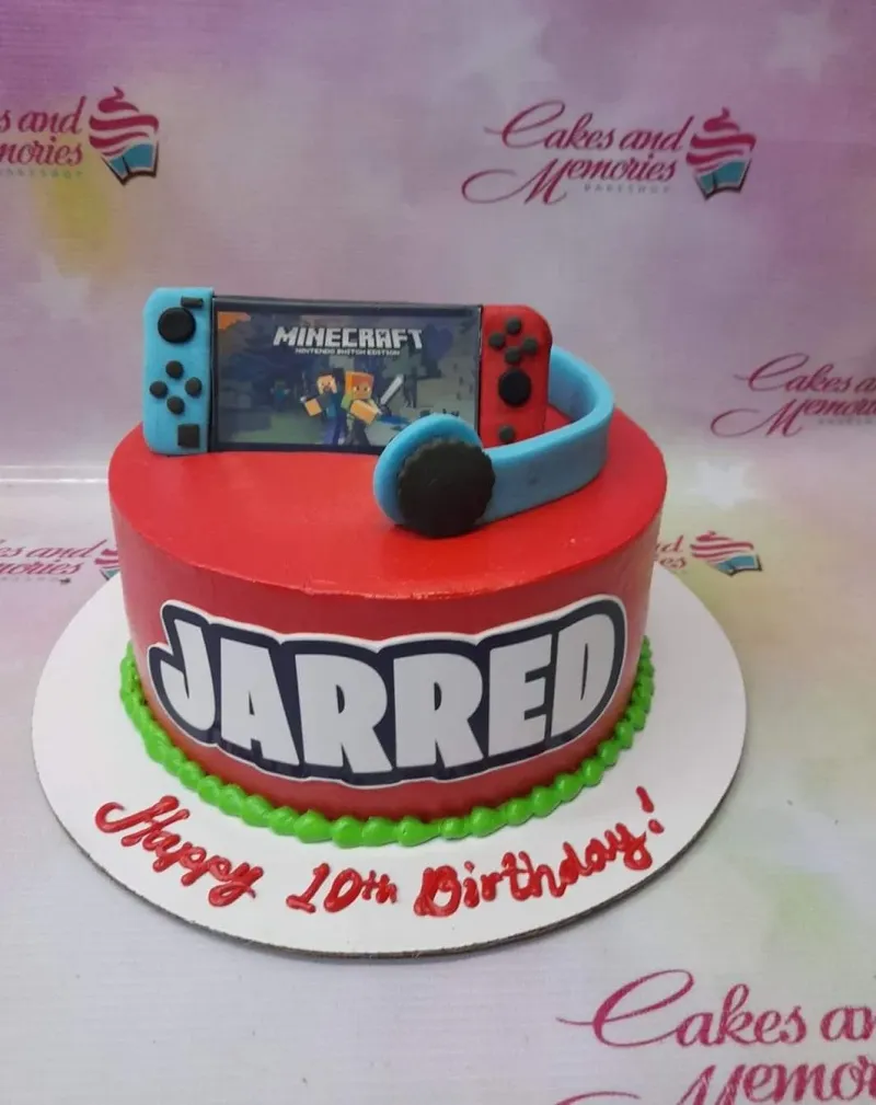 A red 1-tier cake themed Minecraft Nintendo Switch, featuring a Nintendo Switch console printout, 'Jarred' name cutout, and green icing border. A red 1-tier cake themed Minecraft Nintendo Switch, featuring a Nintendo Switch console printout, 'Jarred' name cutout, and green icing border.